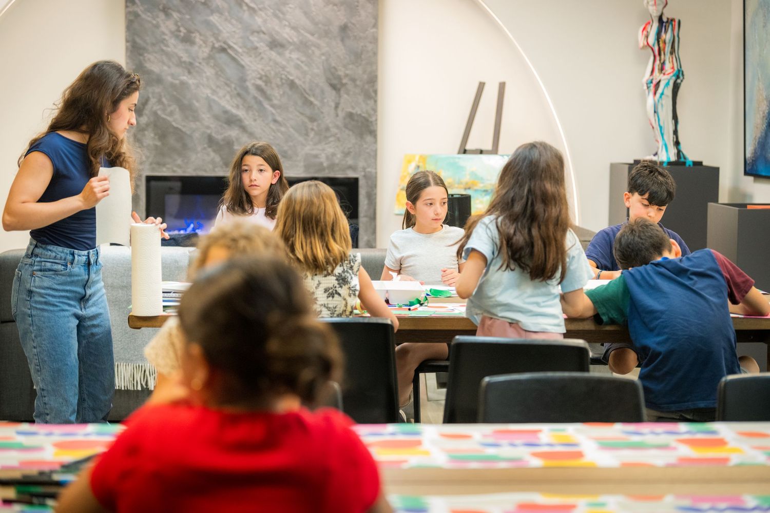 Children and teacher engage in an art class around a large table in a bright, modern studio space.