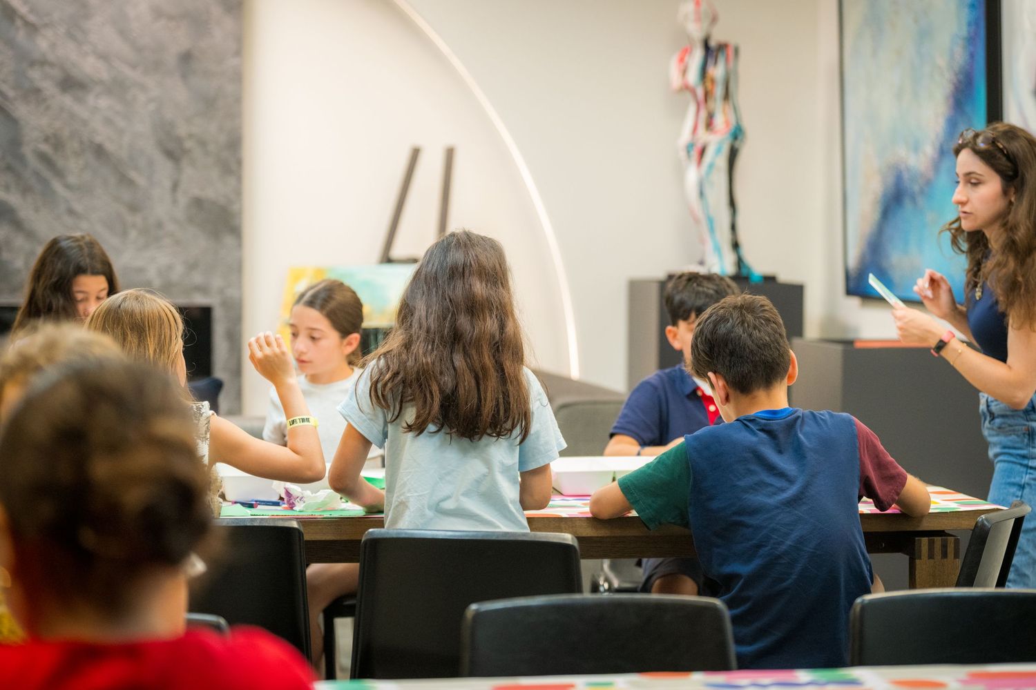 Teacher instructs group of children during an art class in a bright studio with colorful artwork on walls.