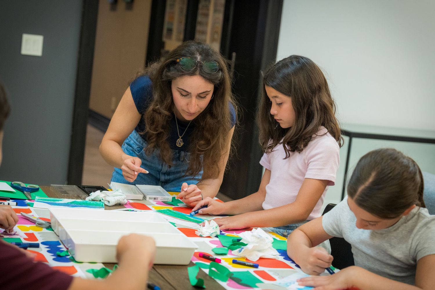 An instructor assists children with a vibrant collage art activity at a table.