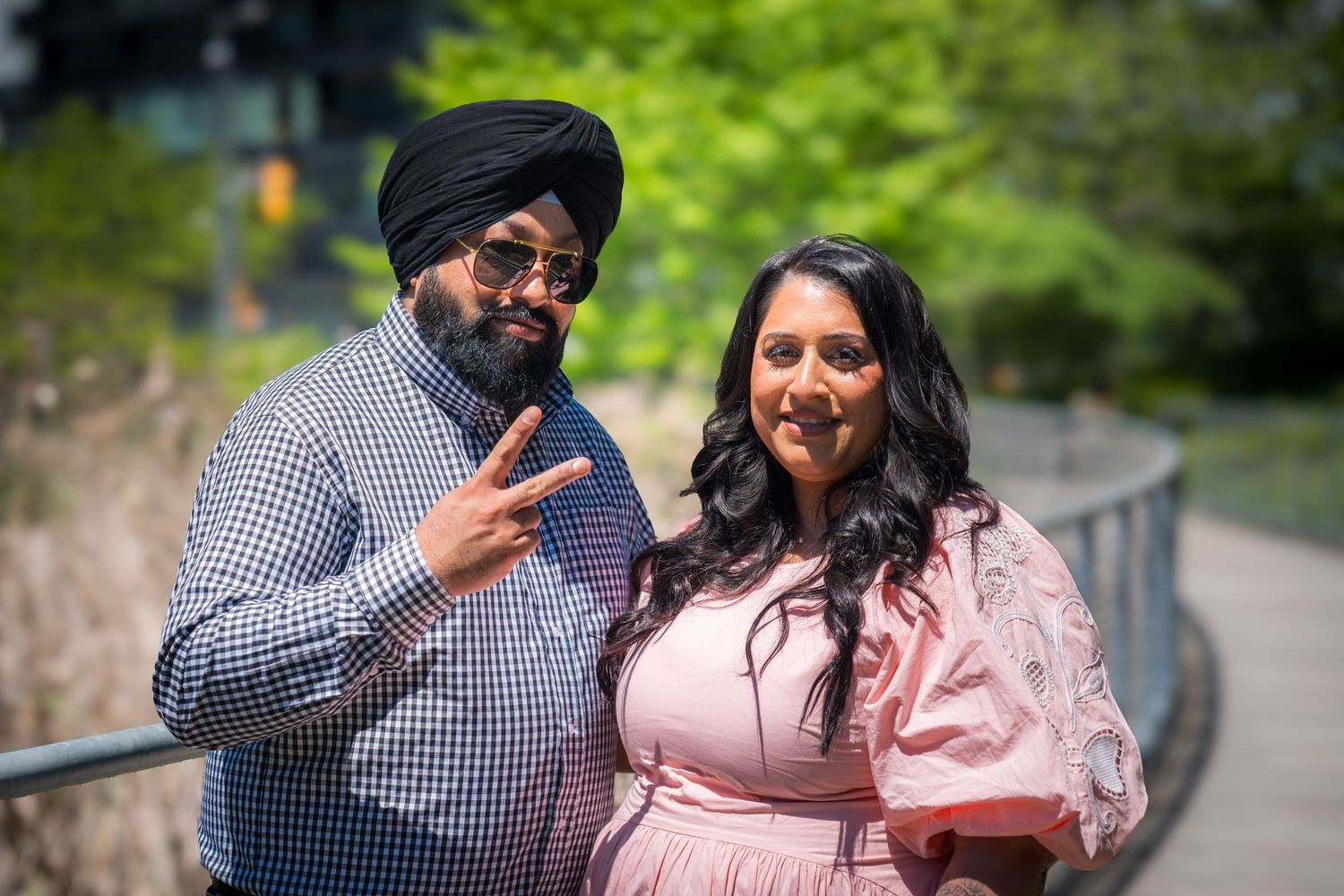 A Sikh man and a woman in pink dress pose outdoors, smiling and making a peace sign near a railing.
