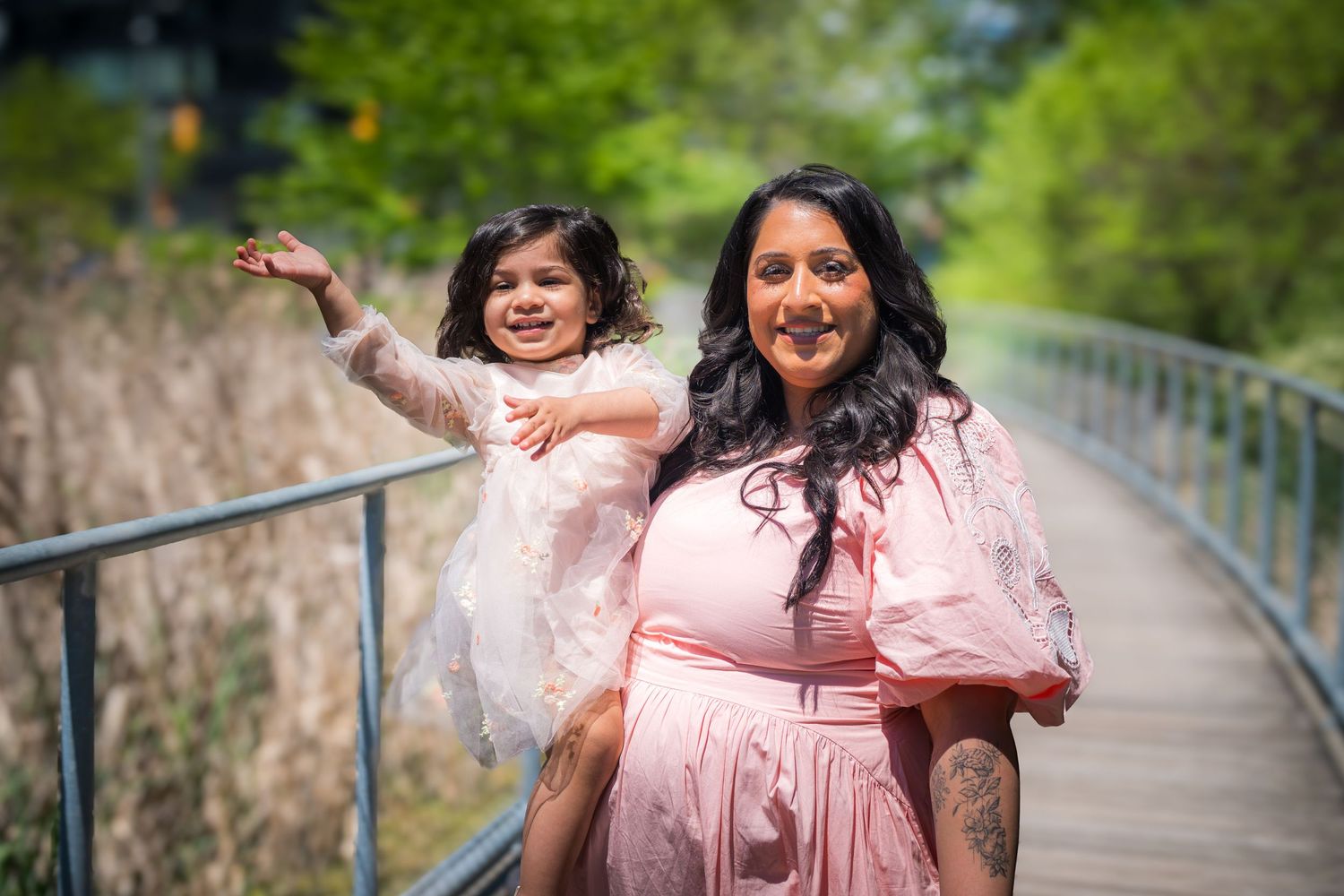 A woman in a pink dress holds a young girl in a white dress on a wooden bridge surrounded by greenery.