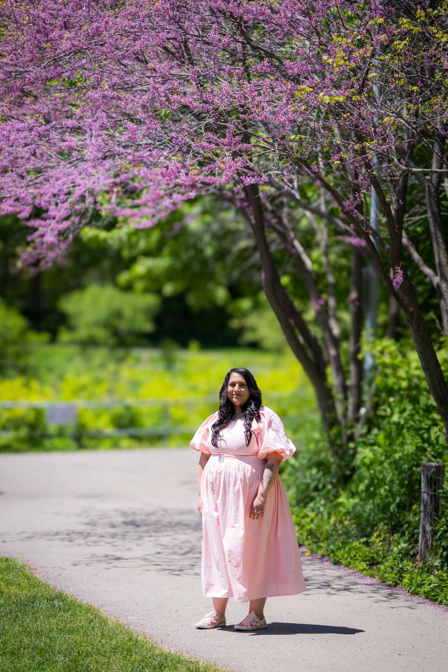 A woman in a flowing pink maternity dress walks under blooming purple redbud trees in a park.
