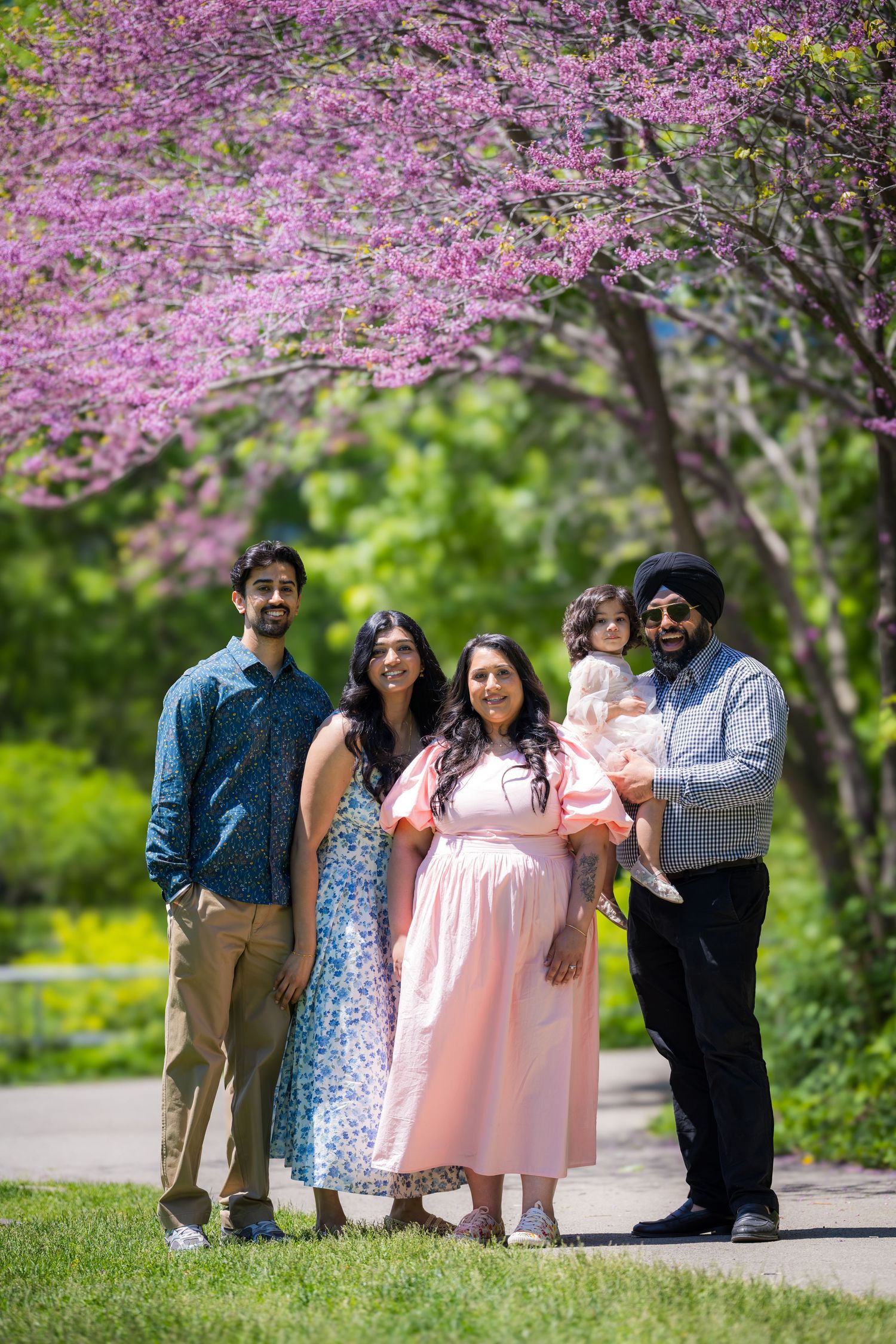 Family of five poses together under blooming pink redbud tree on sunny spring day.