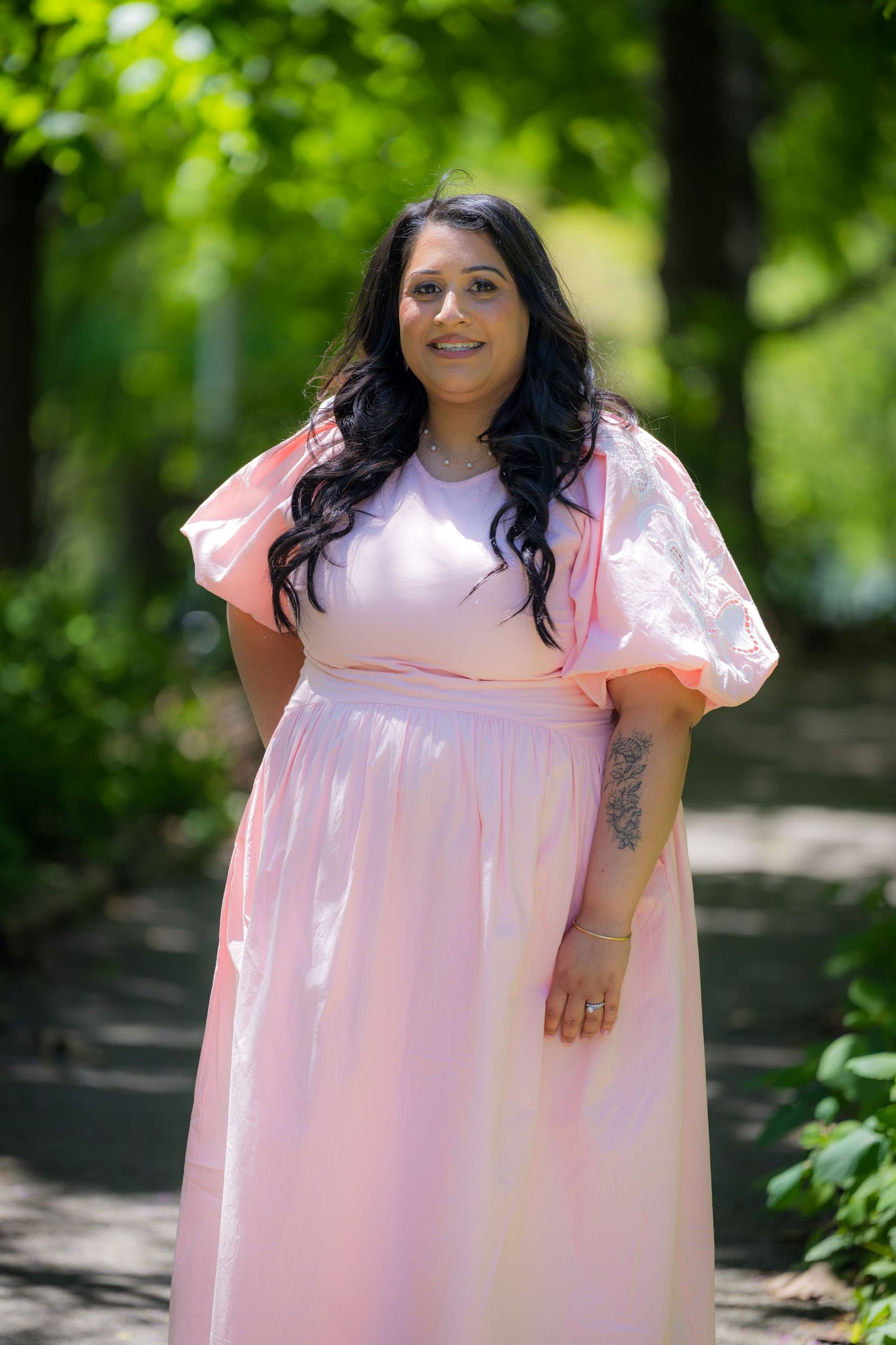 A smiling woman with a tattoo poses in a pink flutter-sleeve maxi dress in a sunny green park.