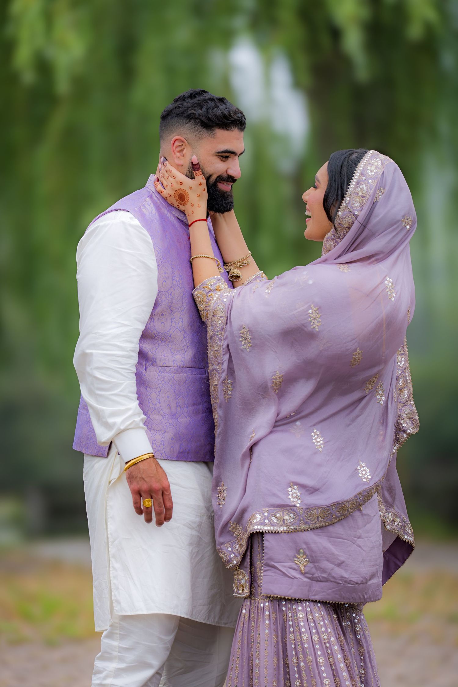 A couple in traditional Indian attire poses playfully under willow trees wearing lavender and white clothing.