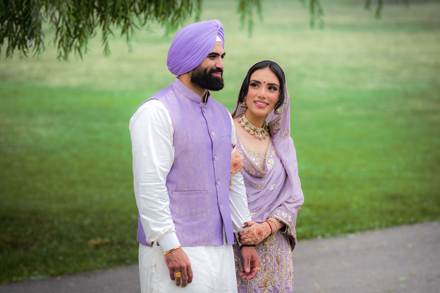 A couple in matching lavender outfits walks together on a garden path framed by willow branches.