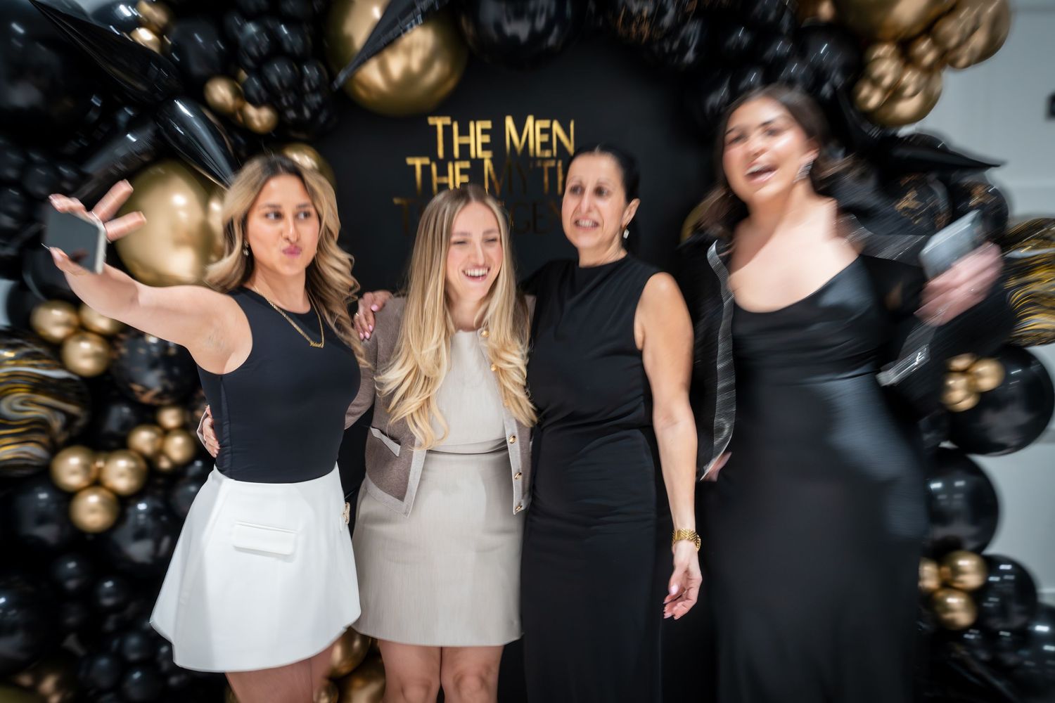 Party guests celebrating in front of black and gold themed event backdrop.
