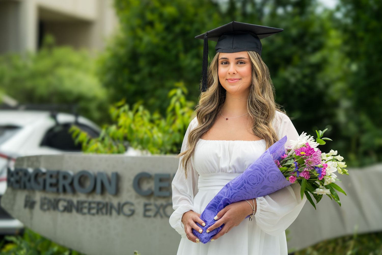 Graduate poses with flowers outside engineering center in white dress and cap.