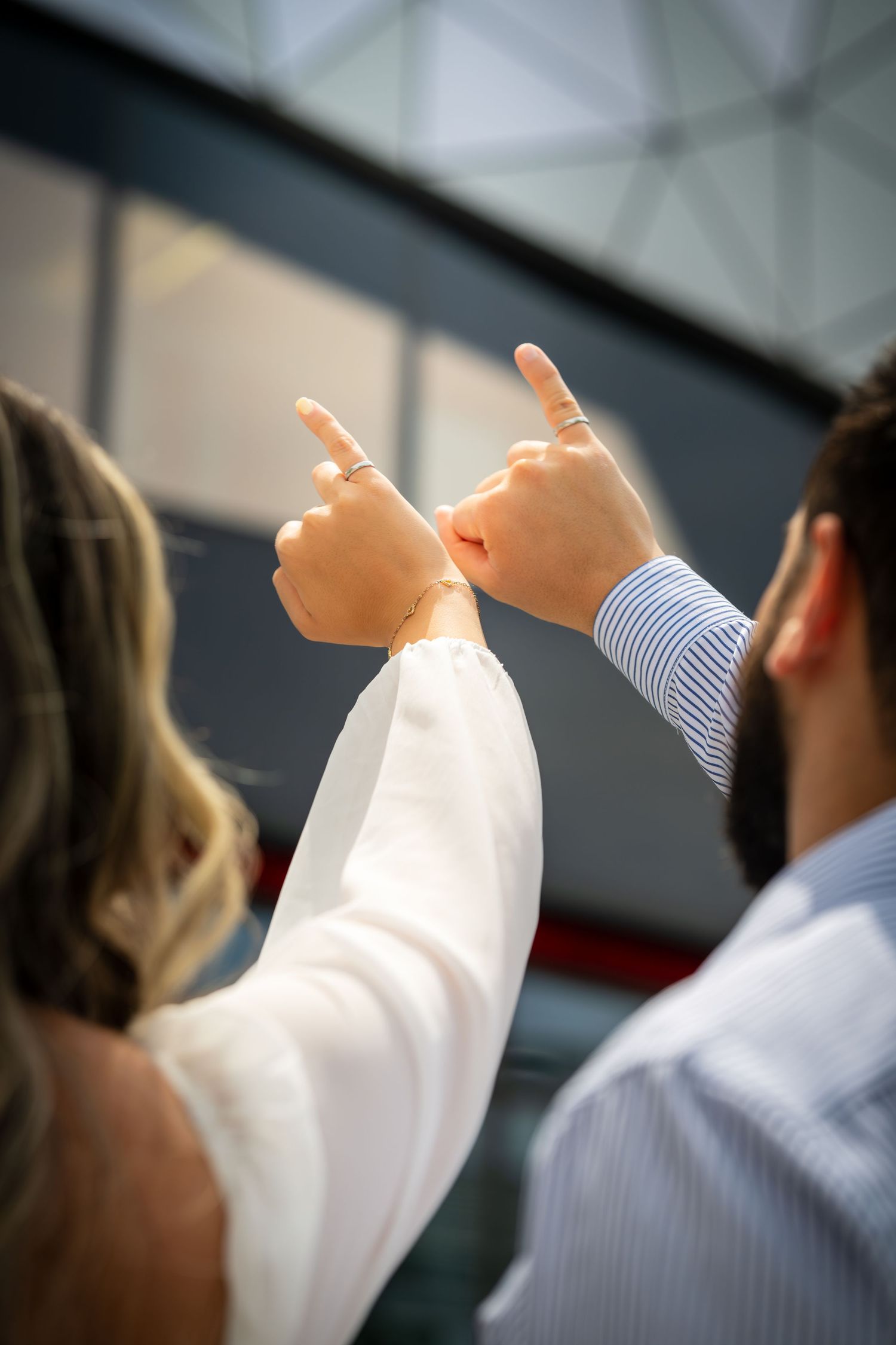 Two people point upward with raised hands against modern building background.