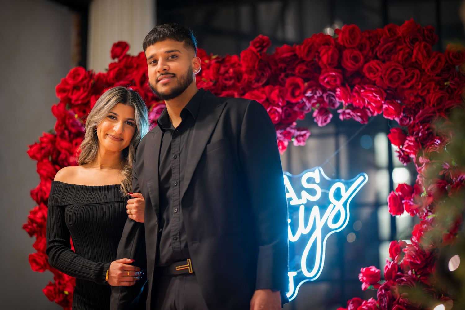 Couple in matching black outfits pose together against red roses and blue neon lighting.