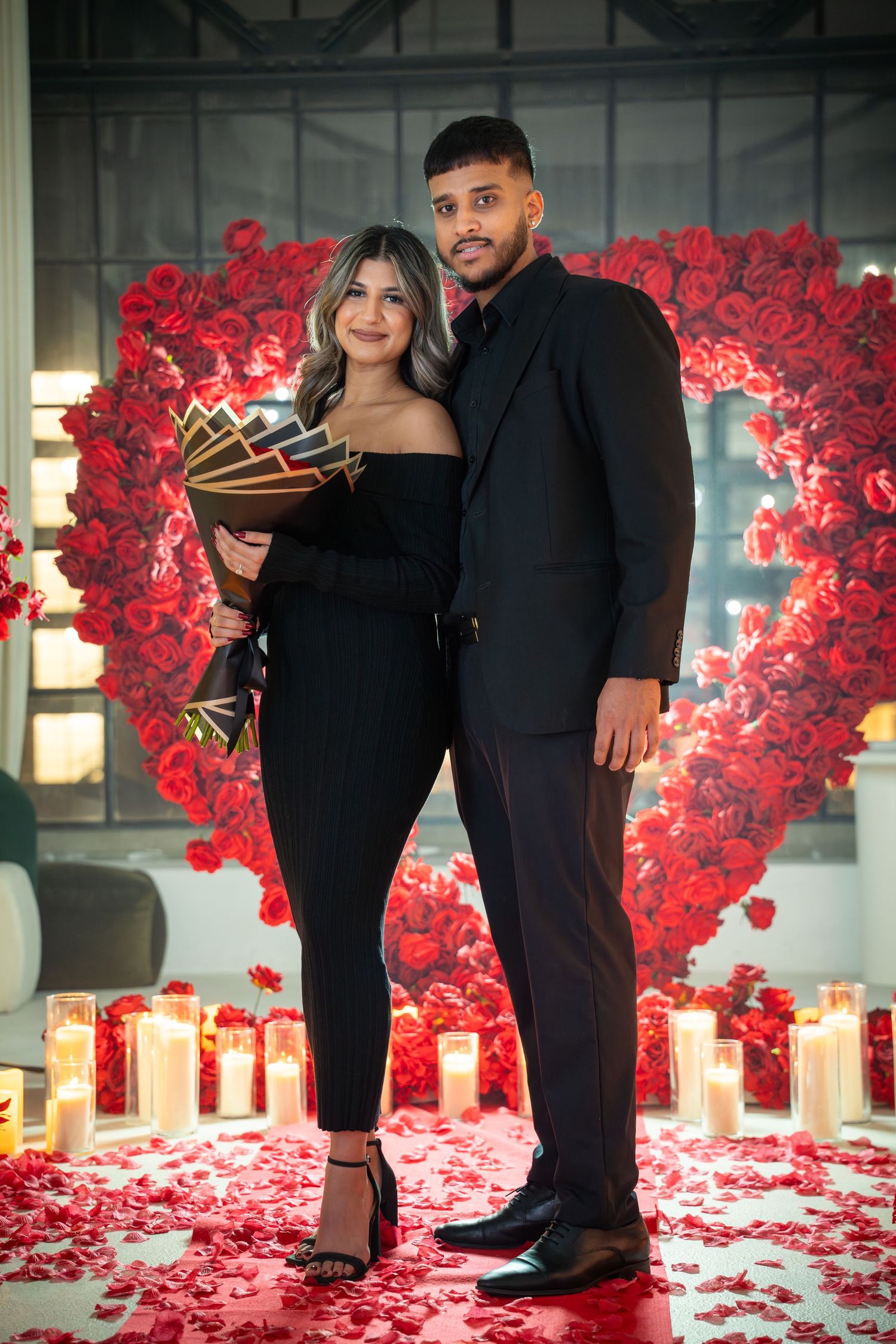 Elegant couple standing among red rose petals and candles during proposal setup.
