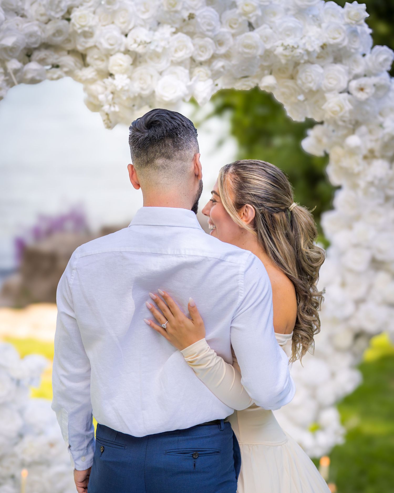 Couple sharing intimate moment under white floral wedding arch with ocean backdrop.
