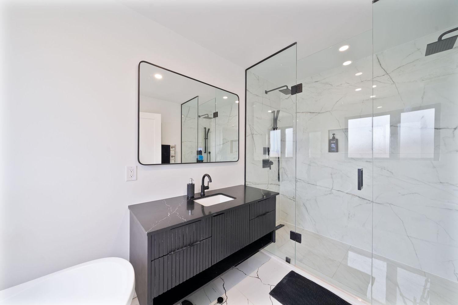 Modern white bathroom interior with black vanity cabinet, frameless glass shower enclosure and large wall mirrors.