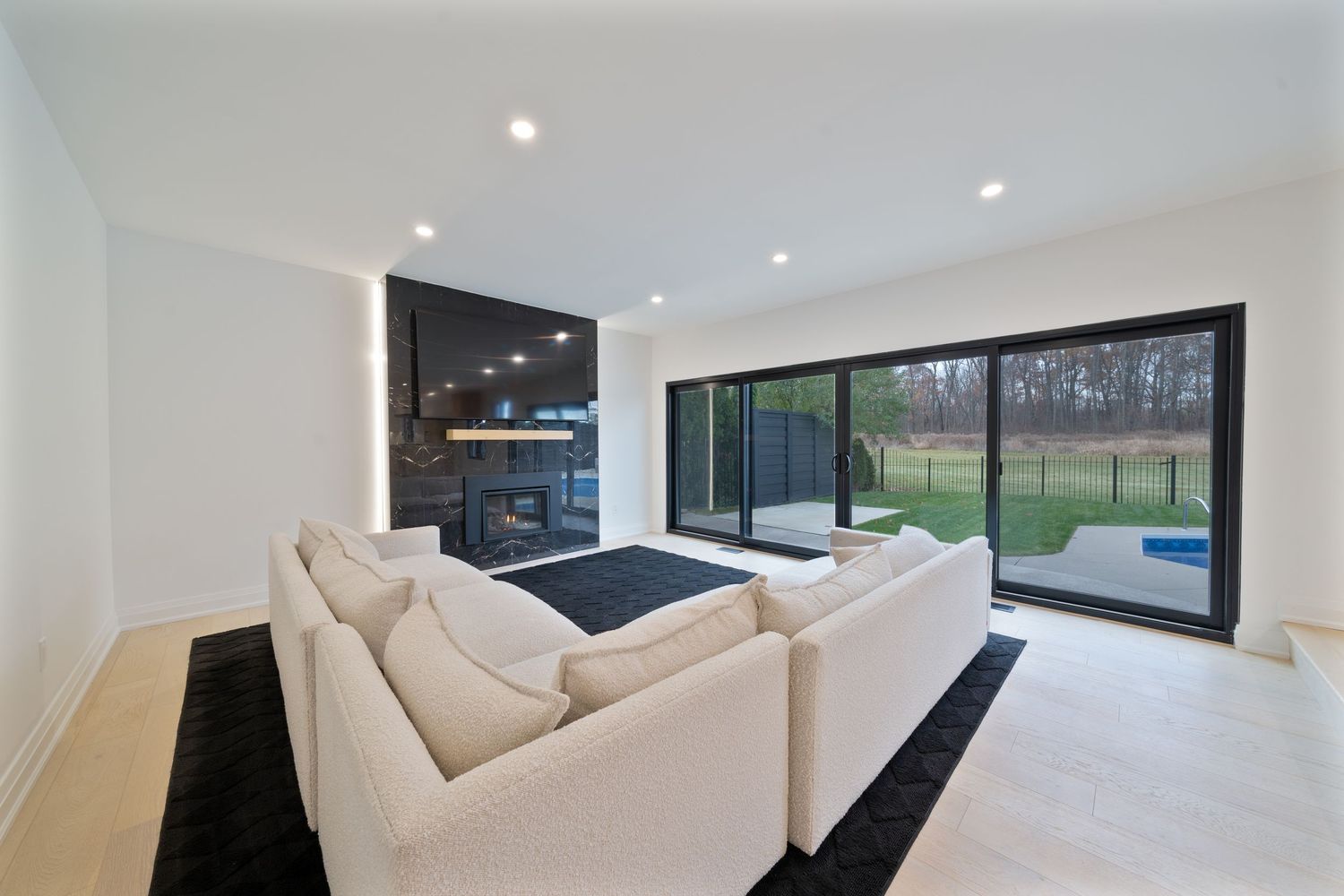 Modern living room with recessed lighting, white walls, and large windows overlooking a green outdoor space.