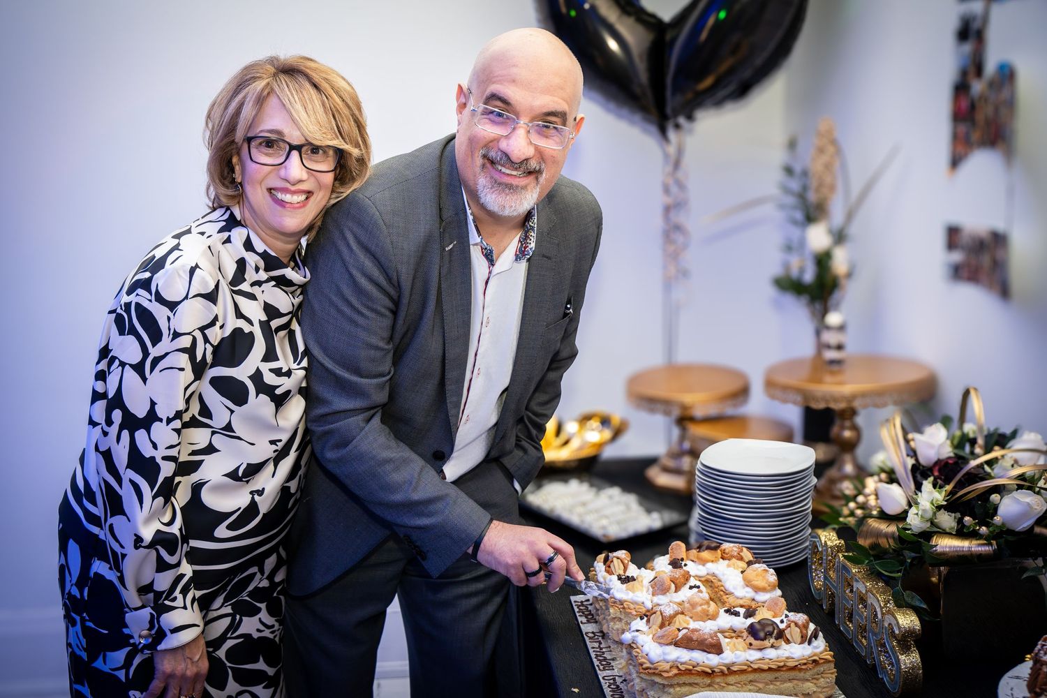 A couple poses together by a dessert table with party decorations in the background.