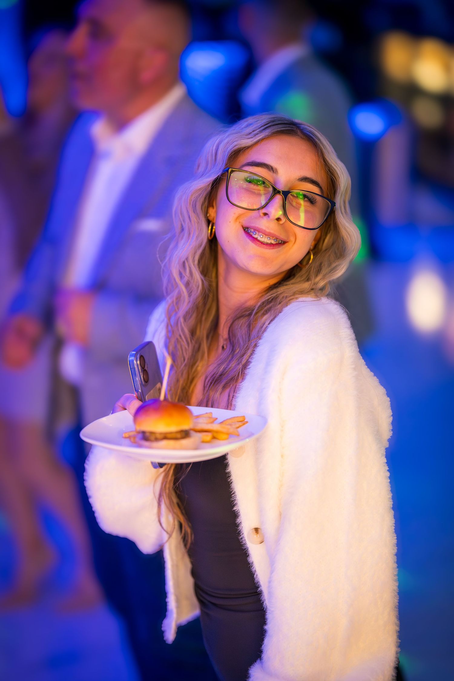 Person in white sweater holding a plate of food at a party with blue ambient lighting.