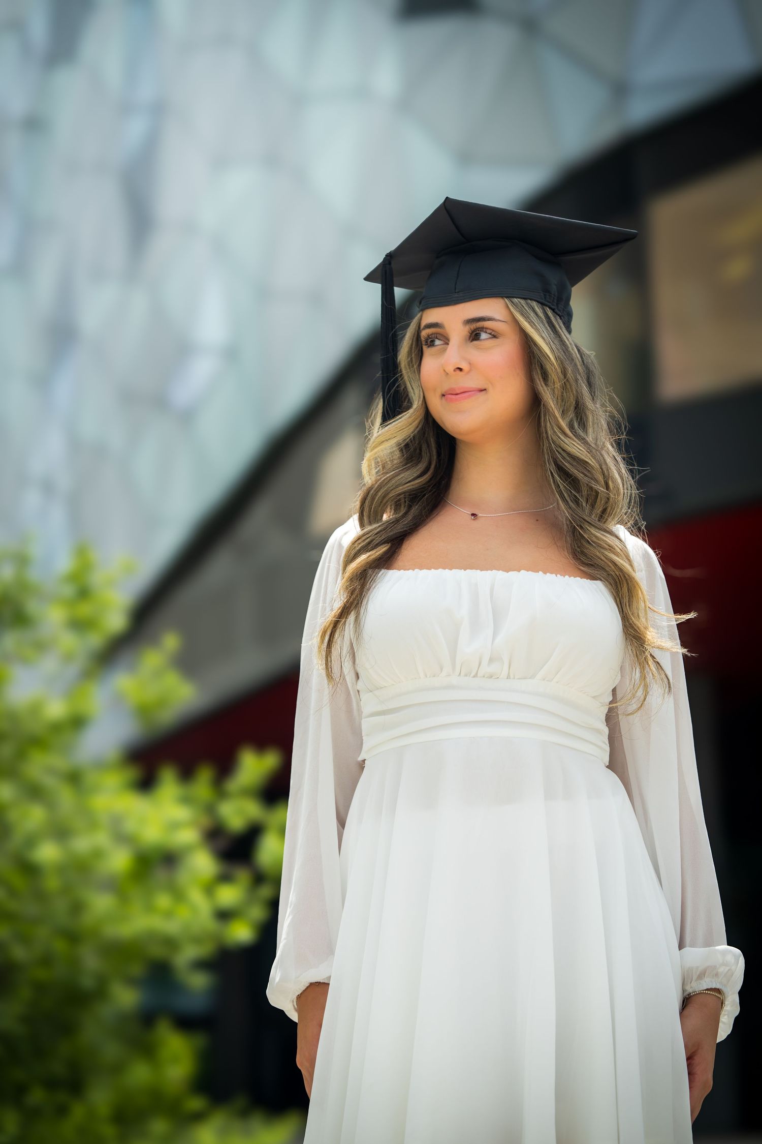 Graduate wearing flowing white dress and mortarboard cap stands in front of stone wall.