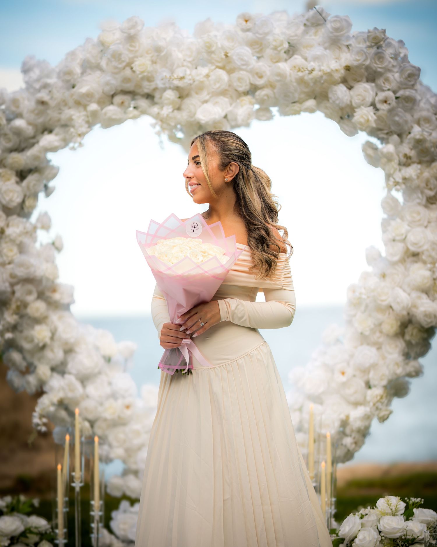 Person poses with bouquet under heart-shaped white floral arch with candles.