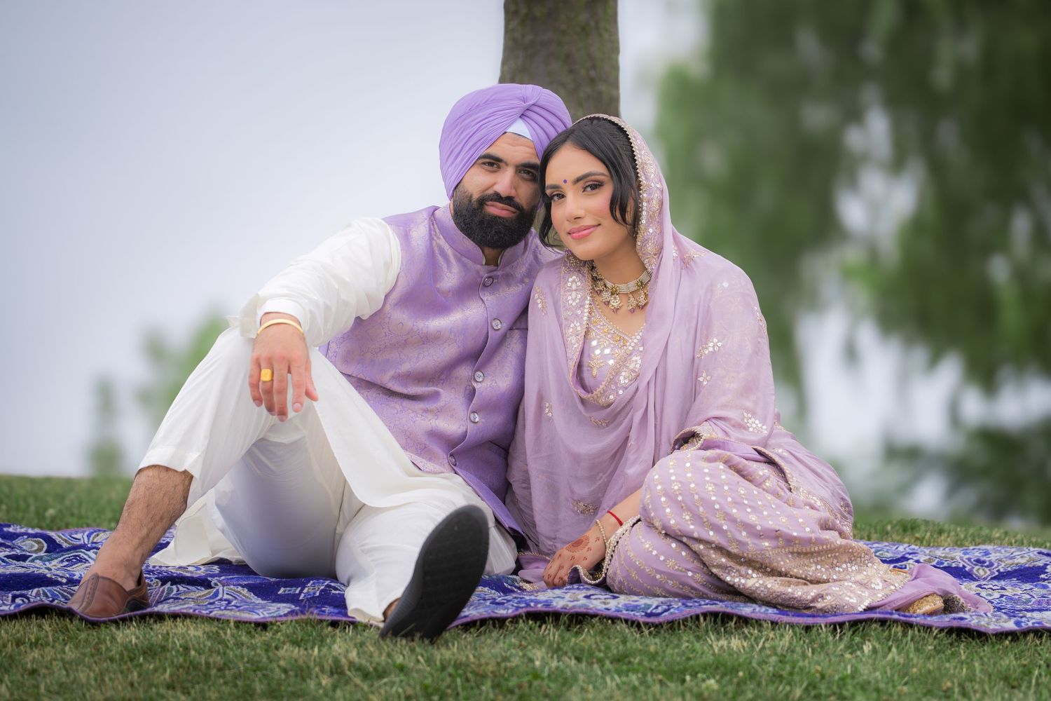 Three people in traditional Punjabi attire relax together on a blue patterned blanket in a park setting.
