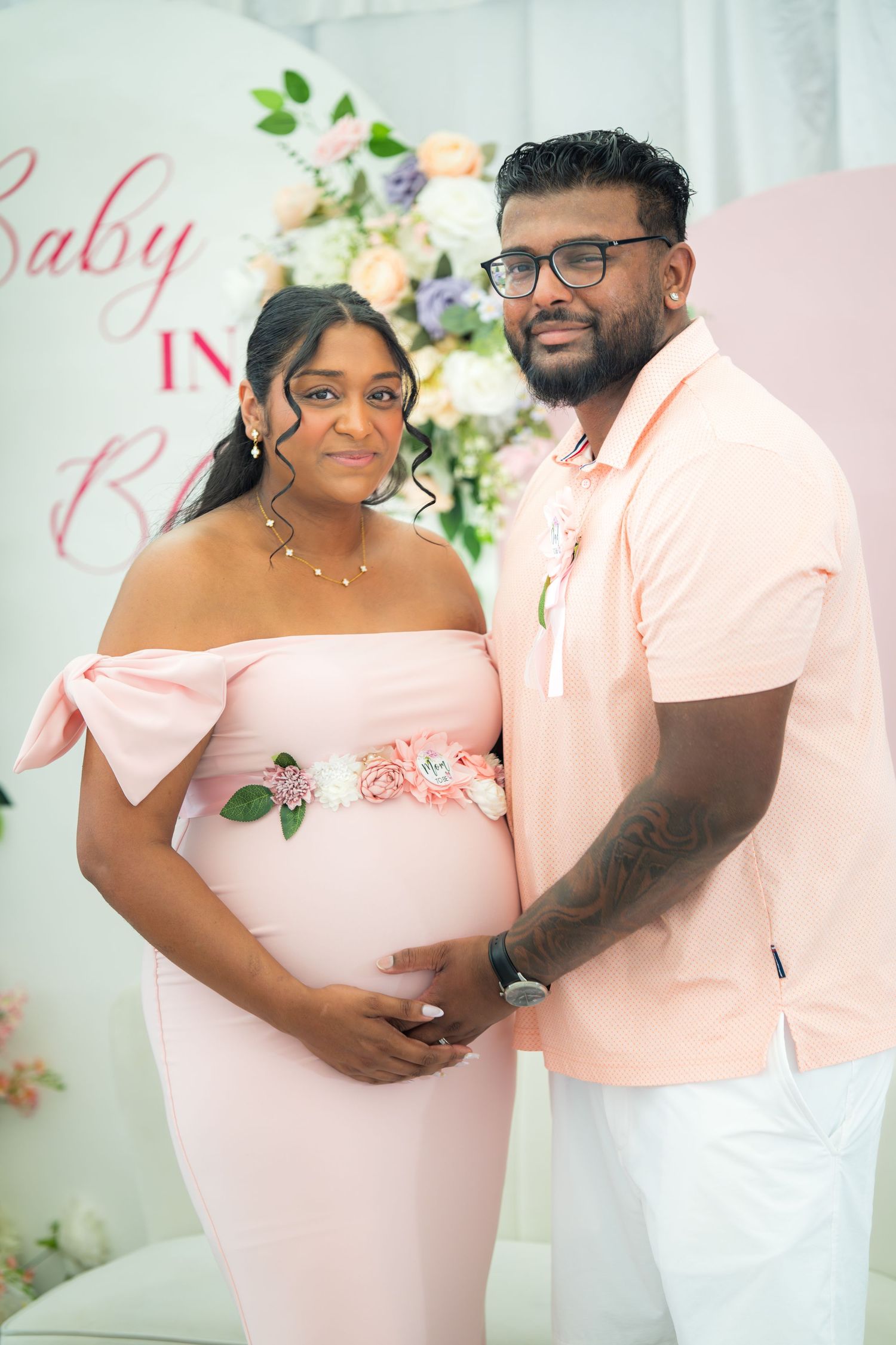 A couple poses in coordinating pink and white outfits against a floral backdrop at a baby-related celebration.