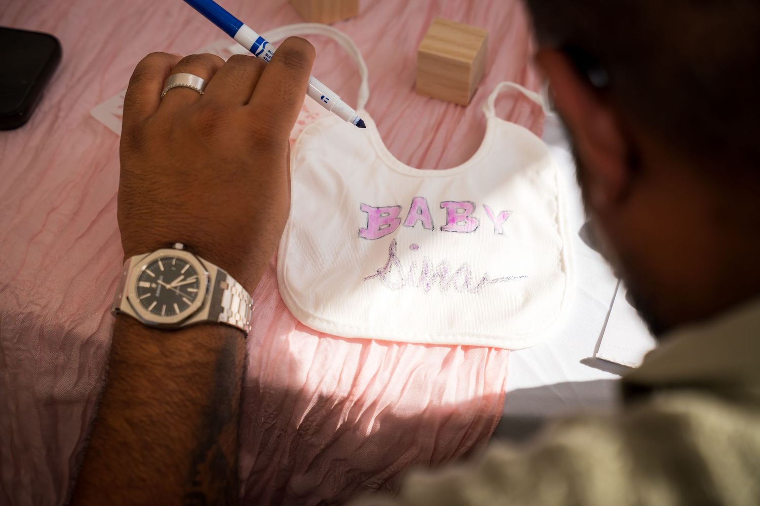 Close-up of hands decorating a white baby bib on pink tablecloth.