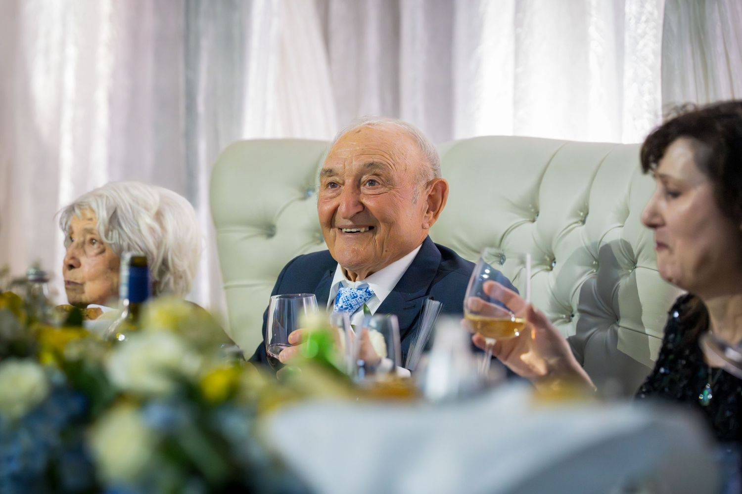Elderly couple smiling and raising glasses at their 65th wedding anniversary party with floral decorations.
