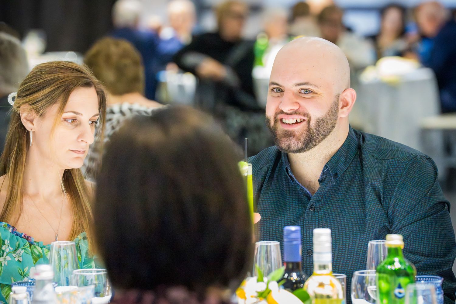 Bald man in black shirt laughing with woman at crowded banquet dinner table.