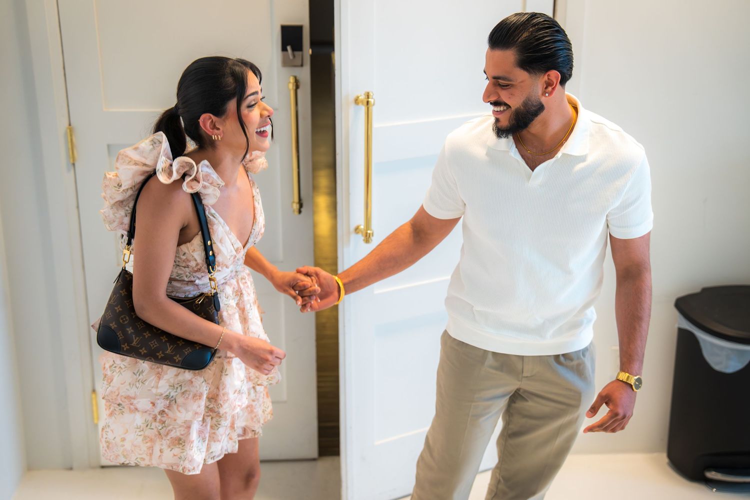A man and woman in elegant attire shake hands and smile warmly in a bright modern room.