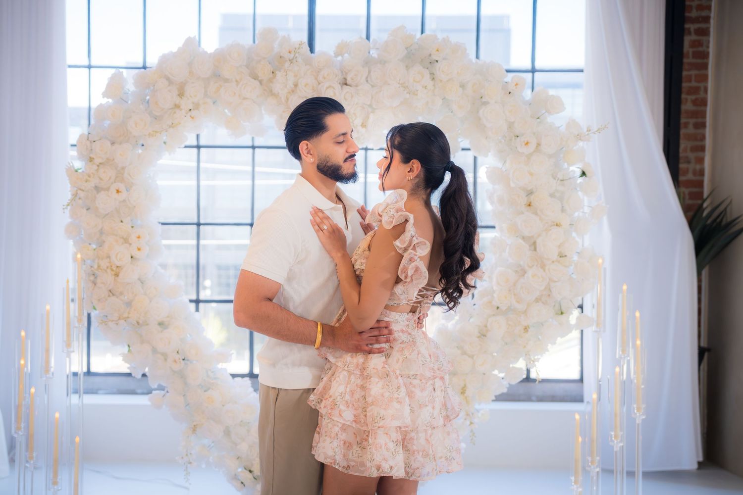 Engaged couple embracing inside a large white floral heart arch in a bright romantic studio.