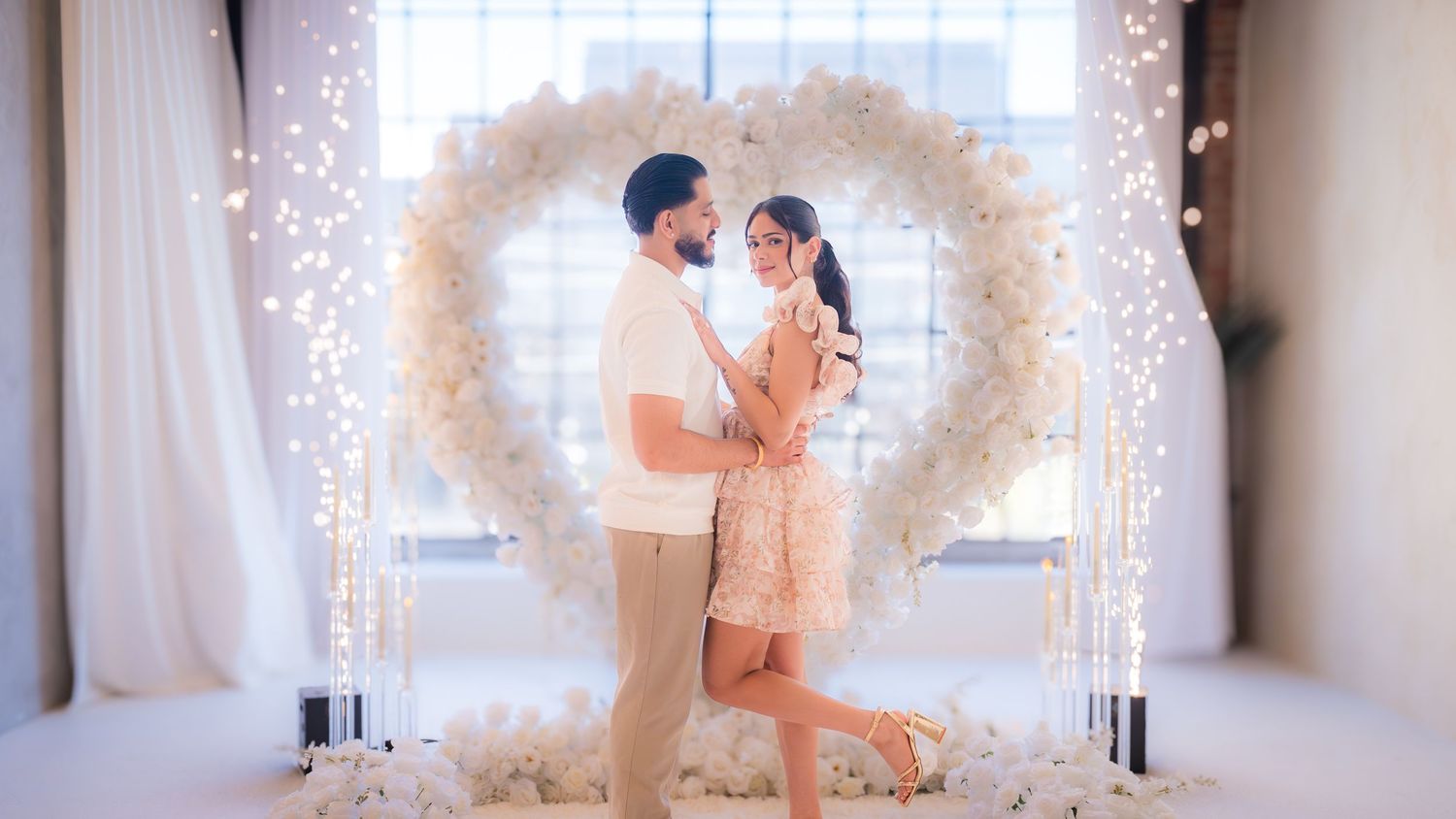 Romantic couple dancing under white heart-shaped floral arch with glowing candles.
