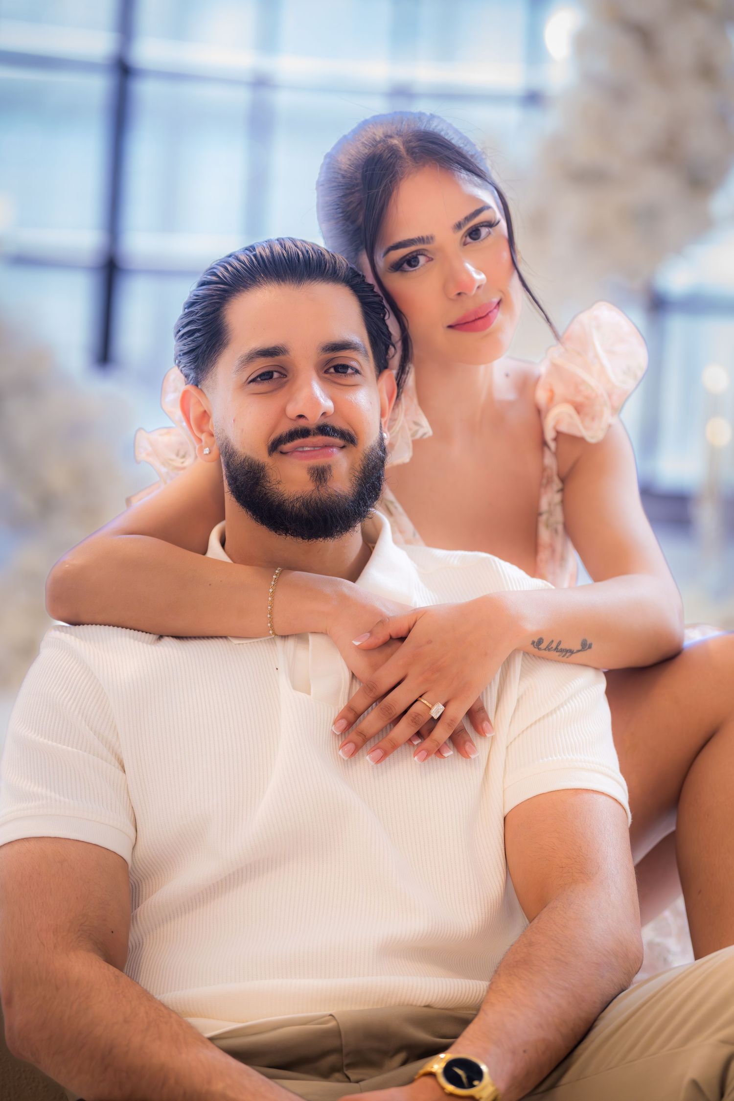 Couple posing for engagement photos, woman in floral dress embracing man in white polo shirt amid white floral decor.