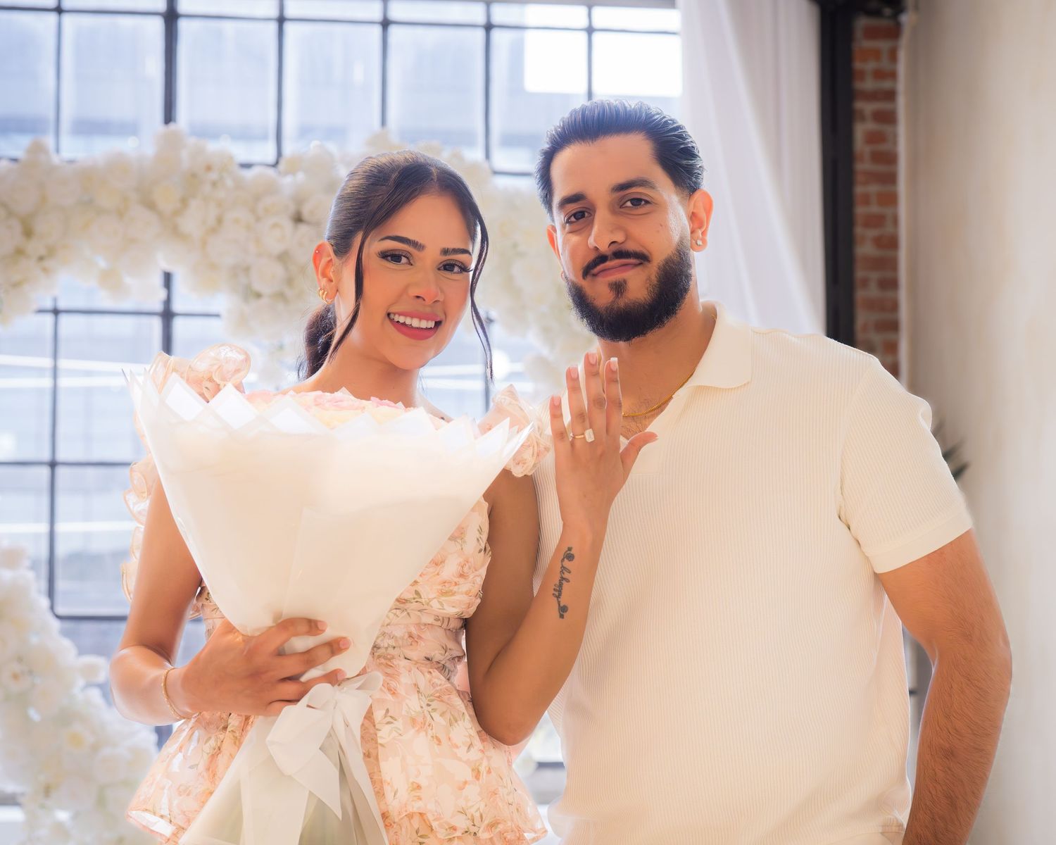 Engaged couple smiling and showing ring while holding white bouquet by floral arch.