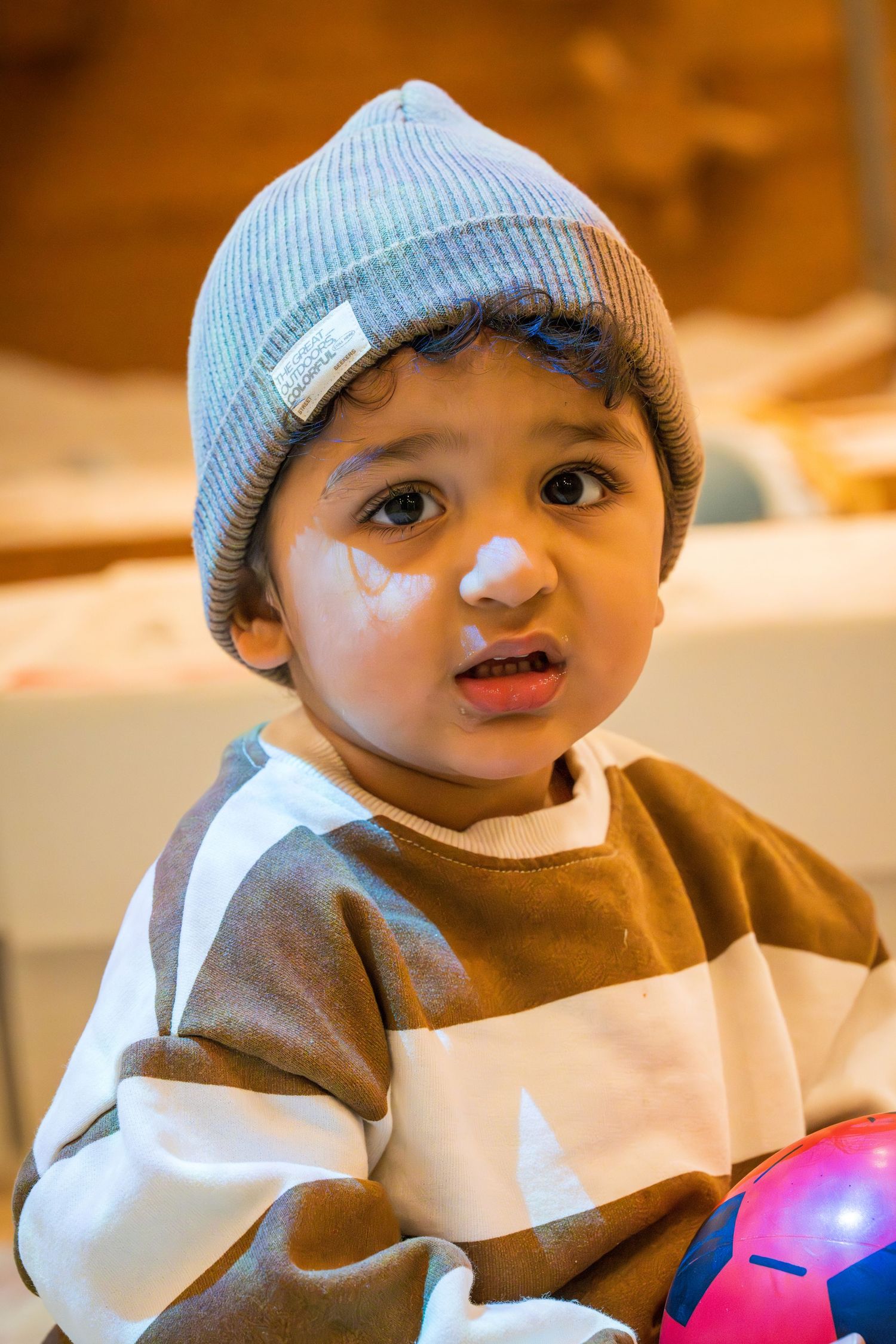 Adorable toddler boy wearing a gray beanie hat and striped brown sweater holding a colorful ball at a party.