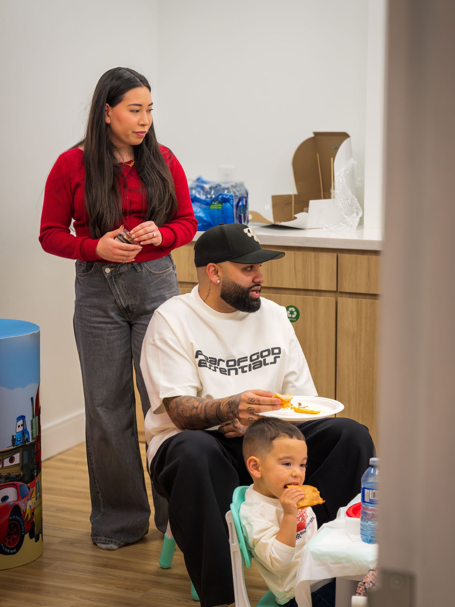 Family at a birthday party with tattooed father in white hoodie eating with young son and woman in red top.