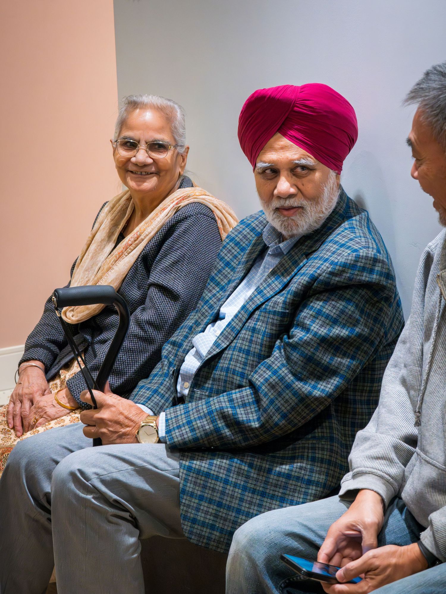 Elderly Sikh man in red turban and plaid blazer sitting with woman and another person in waiting area.