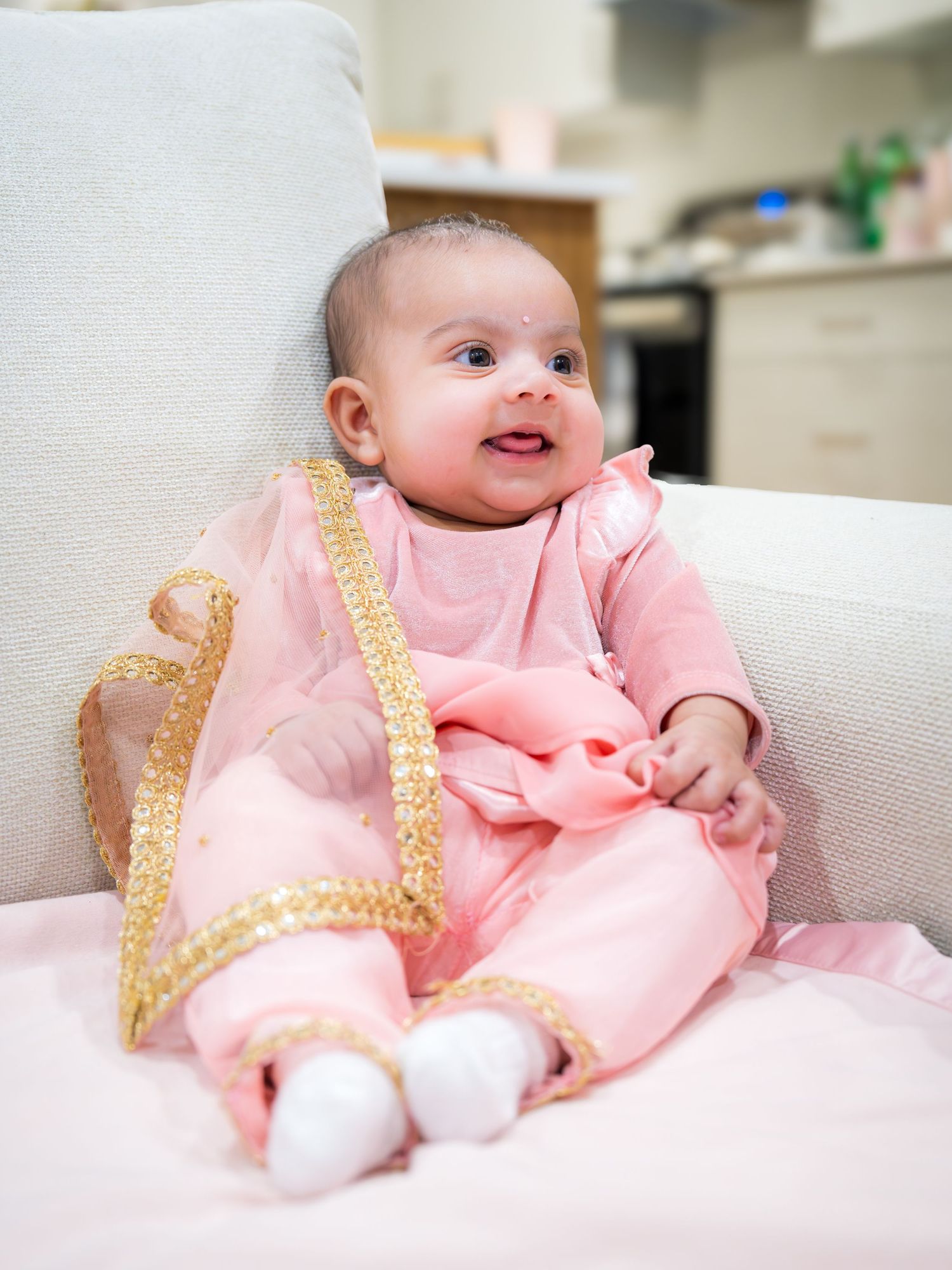 Adorable baby girl in pink and gold Indian outfit sitting on white chair smiling.