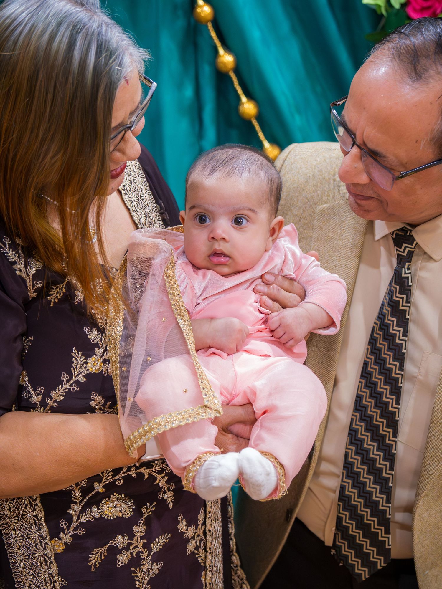 Loving grandparents admiring wide-eyed baby in pink Indian outfit at colorful Lohri celebration.