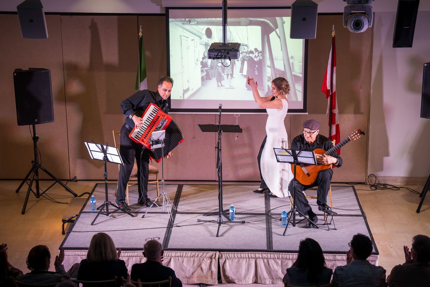 A trio performs on stage with accordion, guitar, and vocalist in white dress before a projected screen.