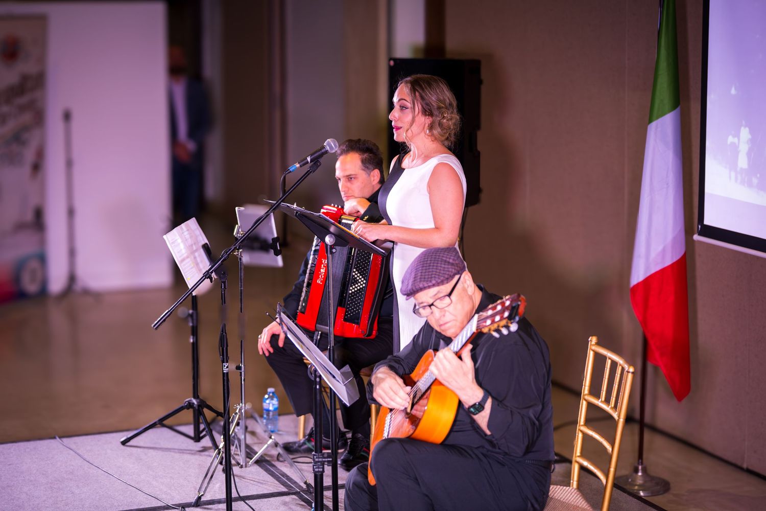 Female singer performs at microphone with accordion and guitar players on stage near an Italian flag.