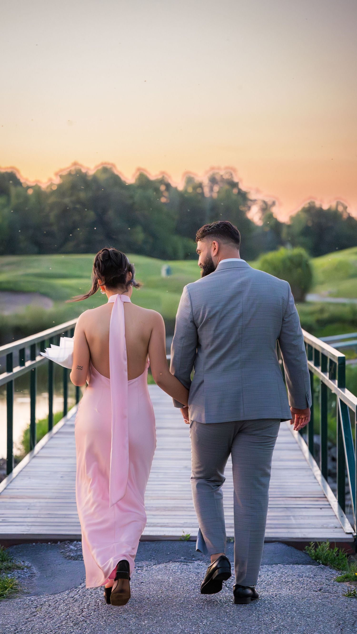 Two people walking along a wooden boardwalk at dusk, one in a pink dress and one in a gray suit.