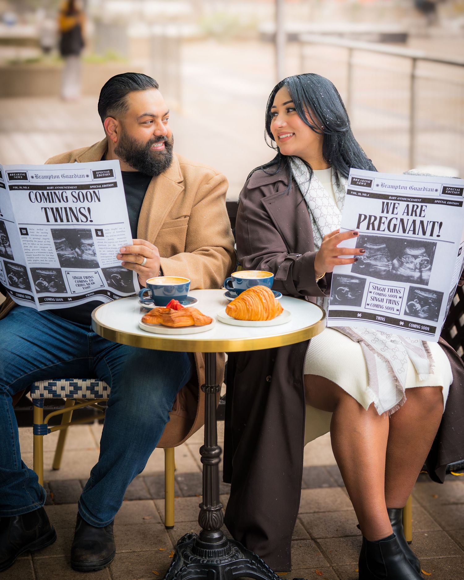 Couple at cafe table holding newspapers with pregnancy announcement headlines.
