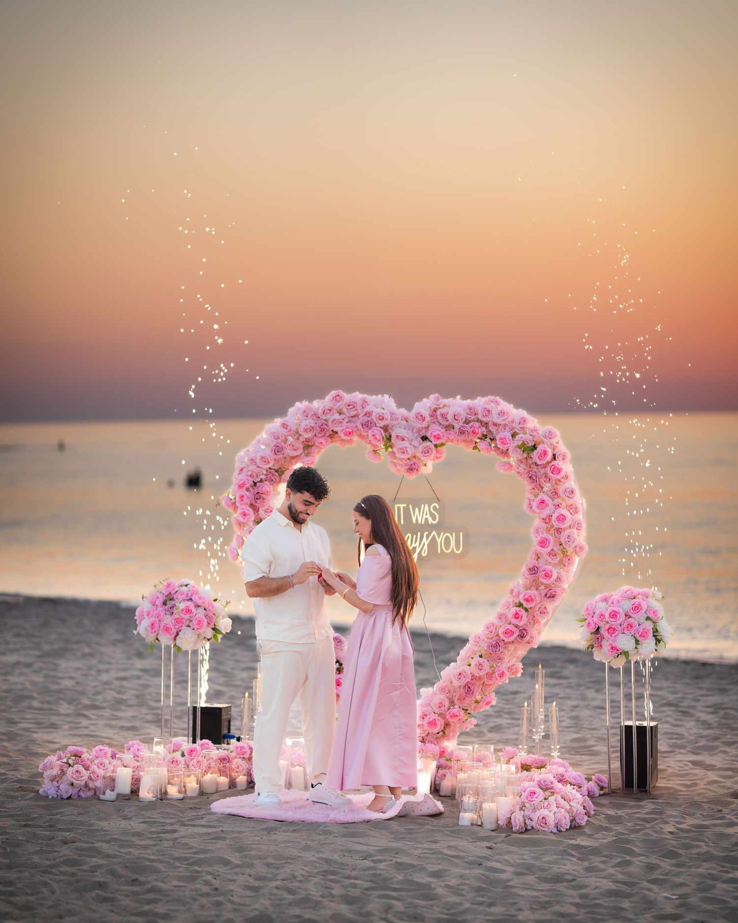 A romantic beach proposal setup with a large pink floral heart arch and candles at sunset.
