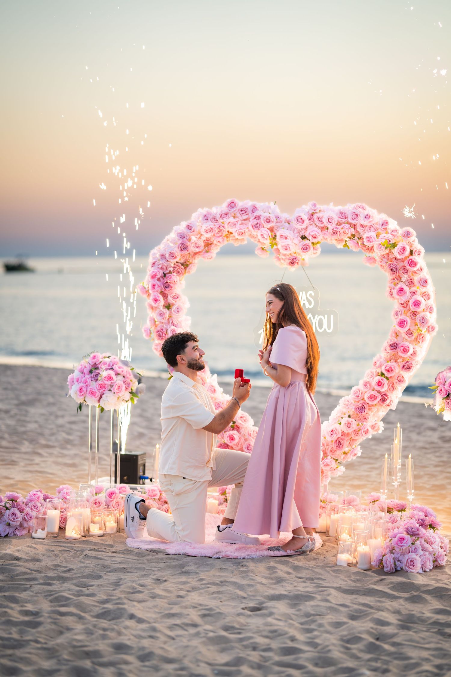 A romantic beach proposal setup with a heart-shaped pink flower arch and glowing candles at sunset by the ocean.