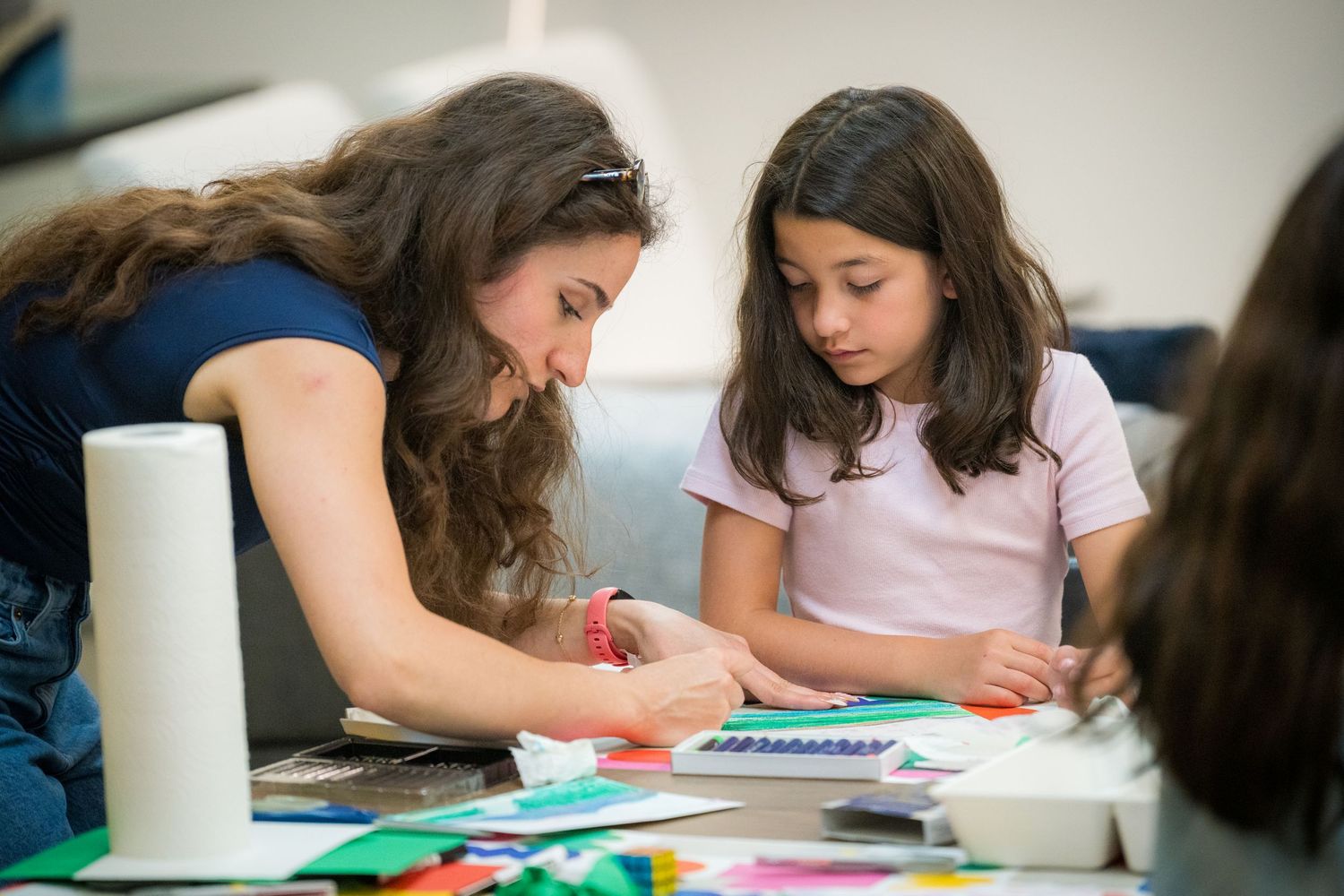 A female teacher assists a young girl with a colorful paper craft project at an art table.