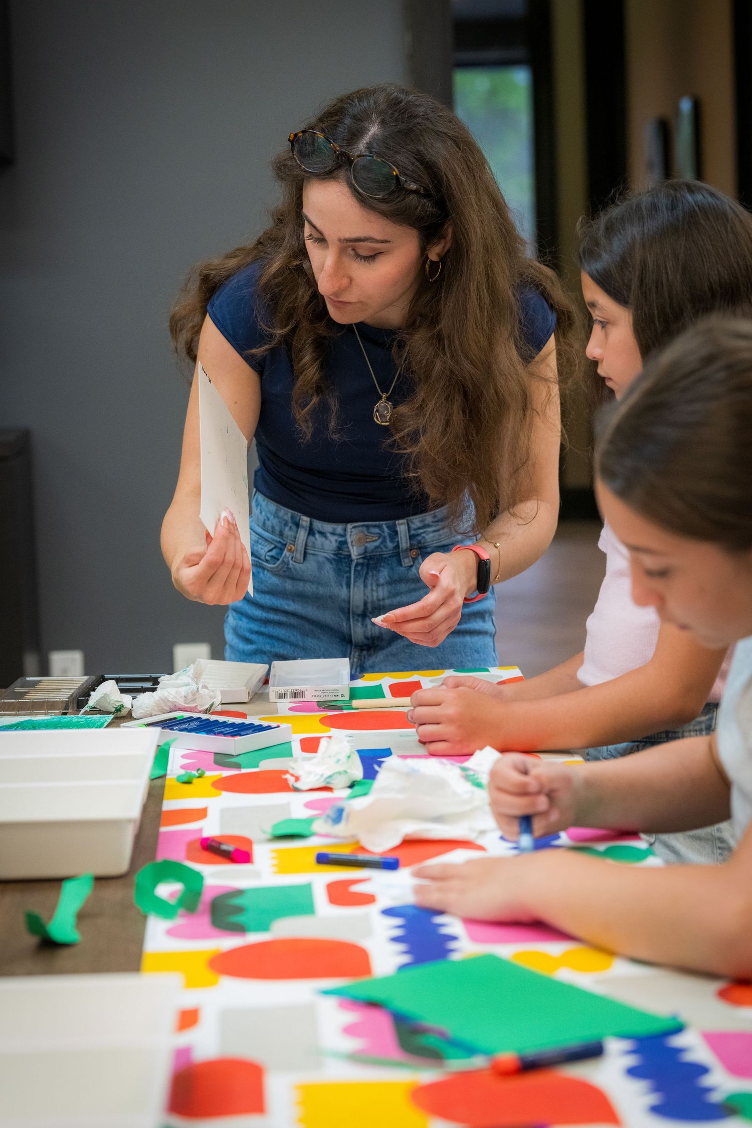 Female art teacher guides two young girls working with colorful paper shapes on a bright table.