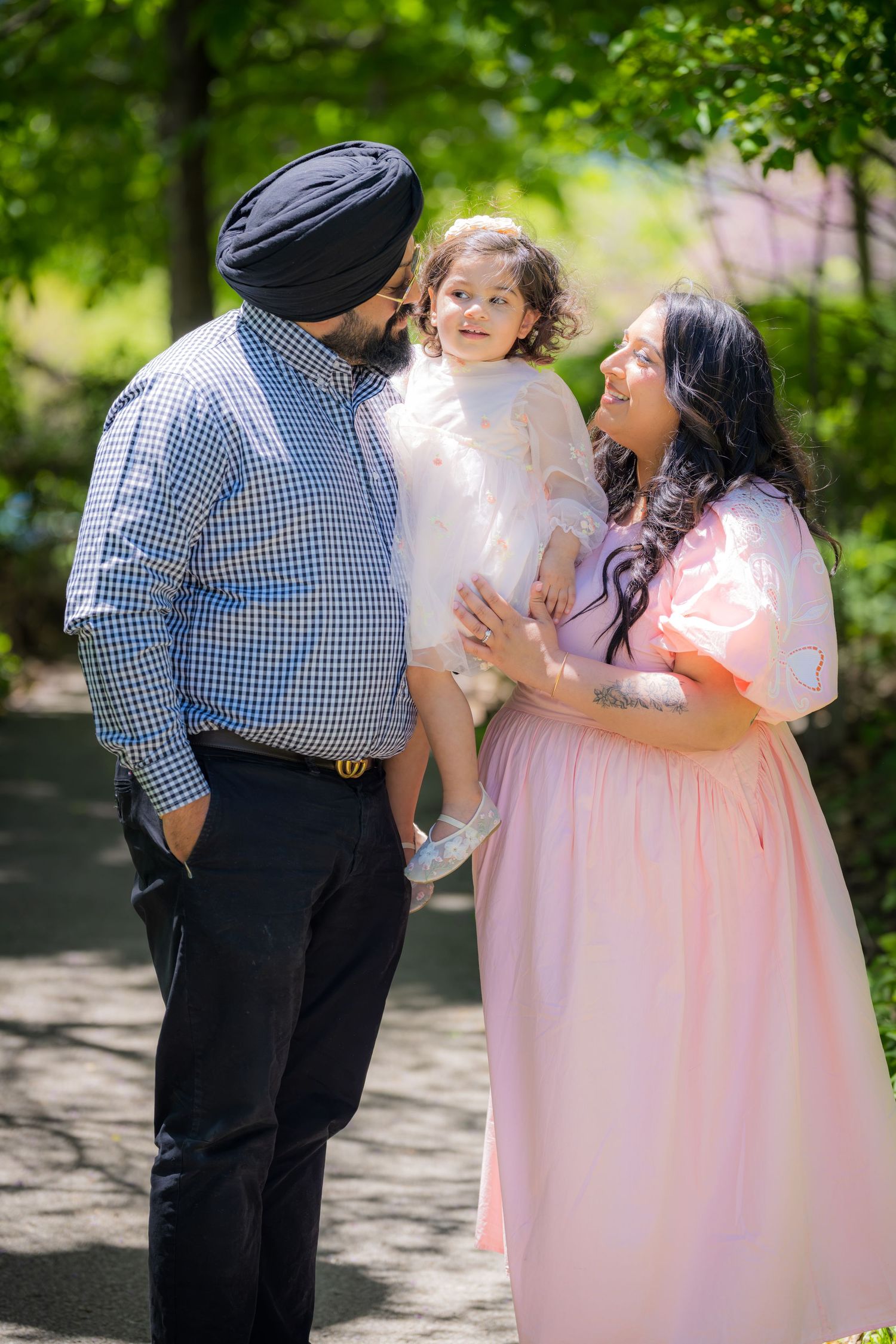 A Sikh family with father in turban and mother in pink dress admire their young daughter outdoors.