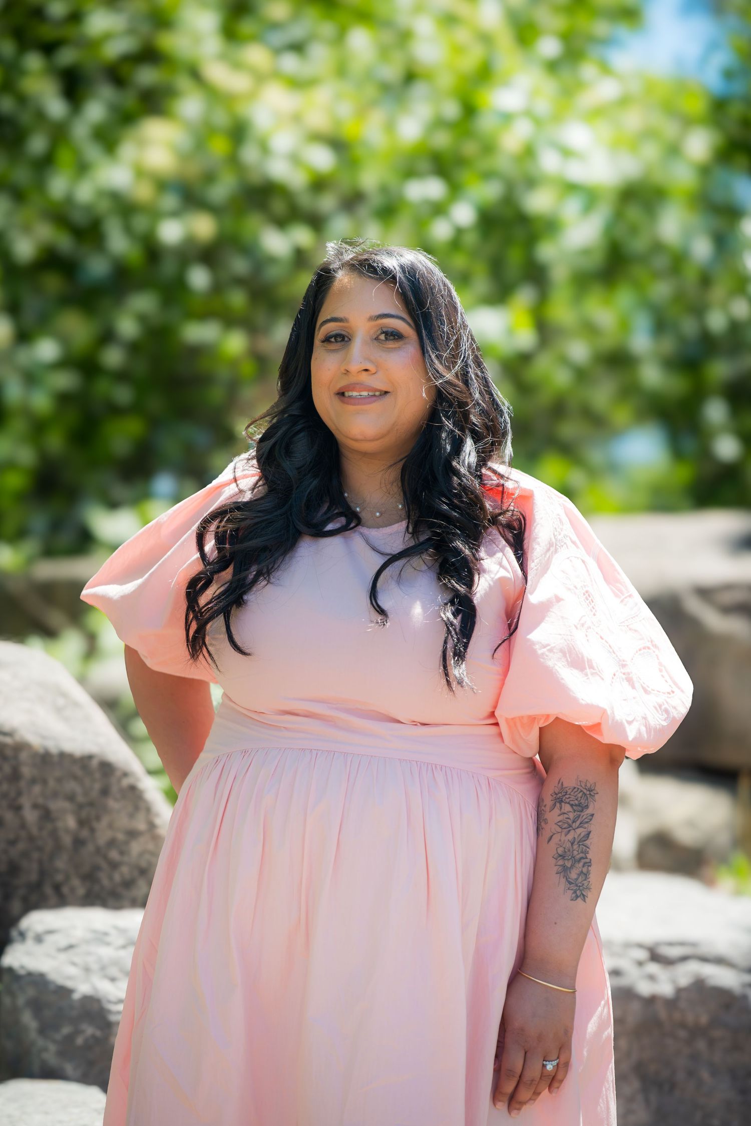 Woman in pink dress with puff sleeves and tattoo smiling outdoors among rocks and greenery.