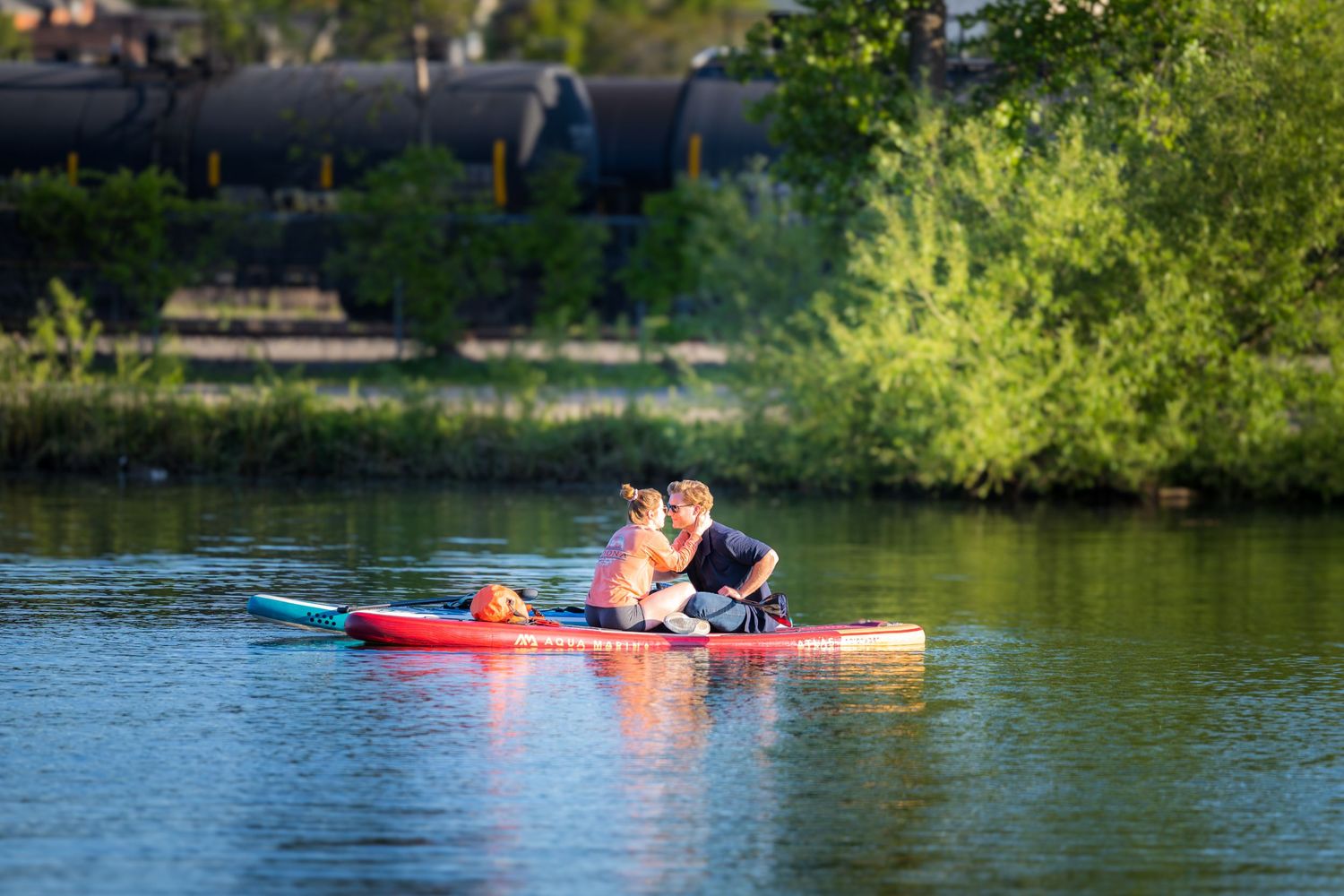 Couple sitting closely together on a red paddleboard paddling on a peaceful river at sunset.