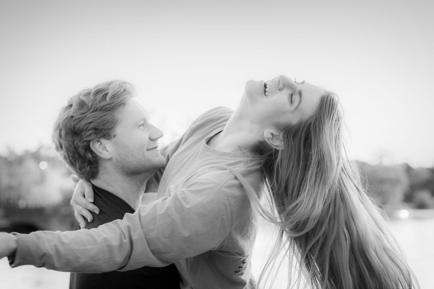 Black and white photo of a joyful couple embracing outdoors, woman laughing with hair flowing in the breeze.