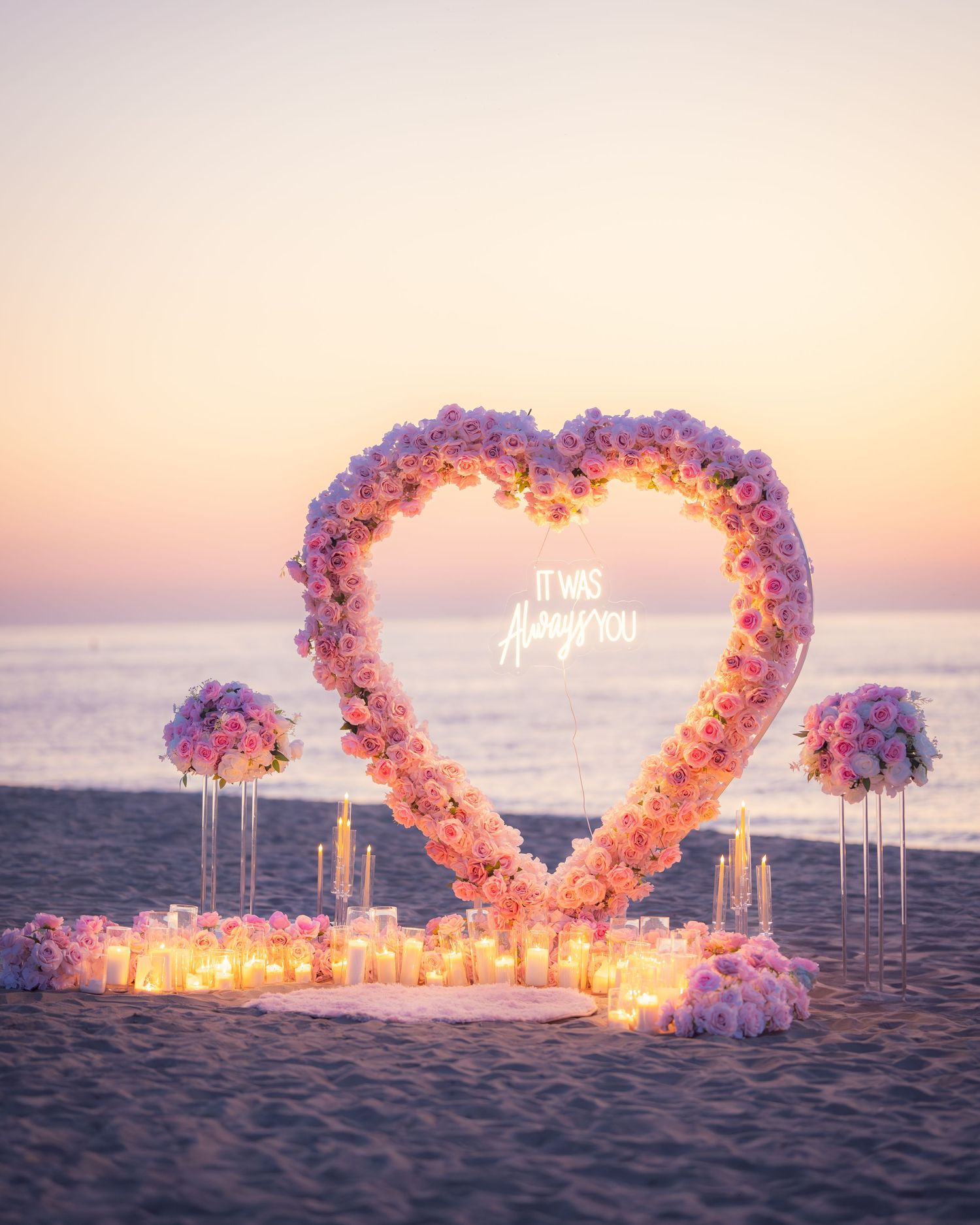 Illuminated heart-shaped floral arch with candles on a beach at sunset creating a romantic seaside setting.